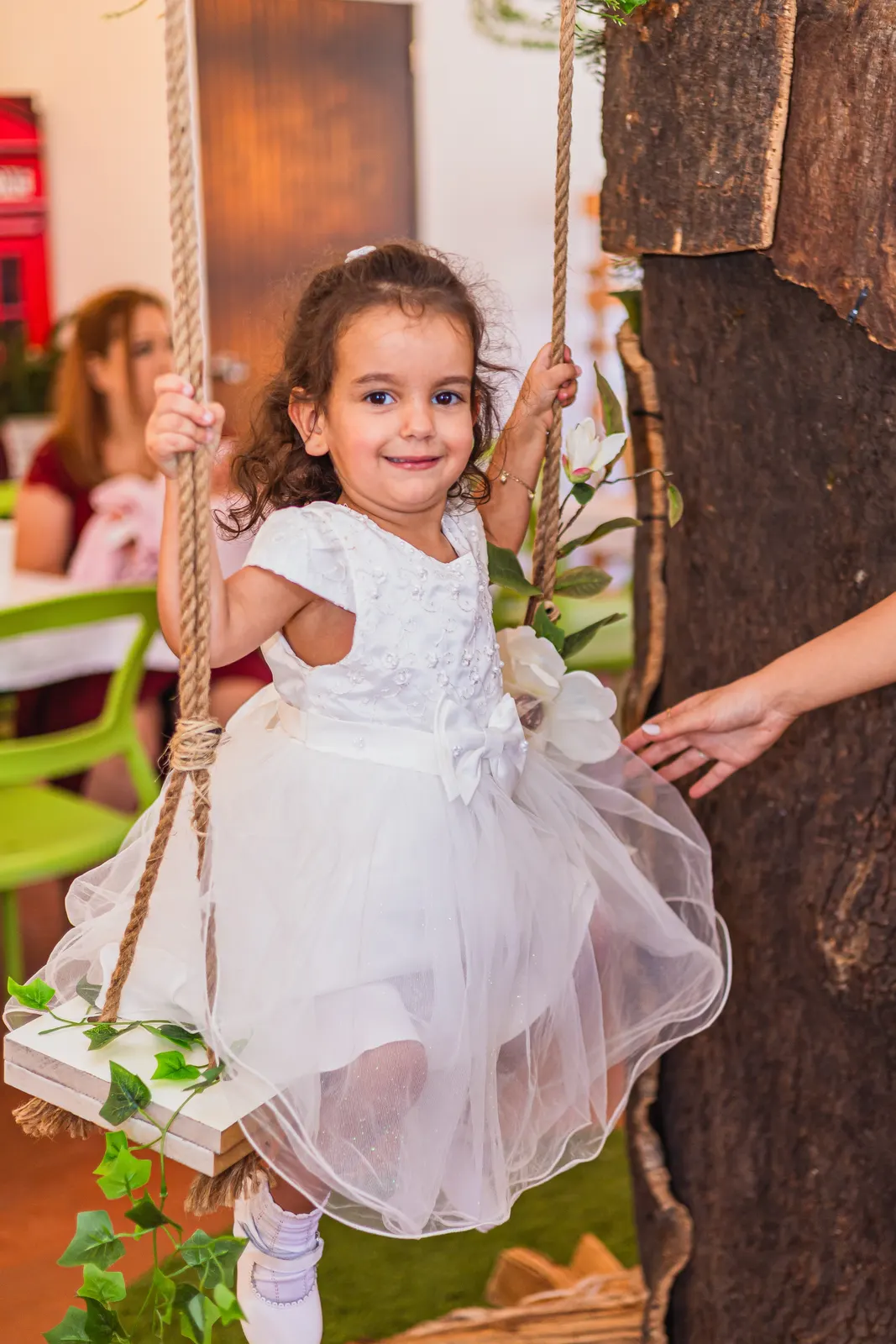 Criança de vestido branco sorri sentada num baloiço de cordas junto a um tronco de árvore decorado com folhagem