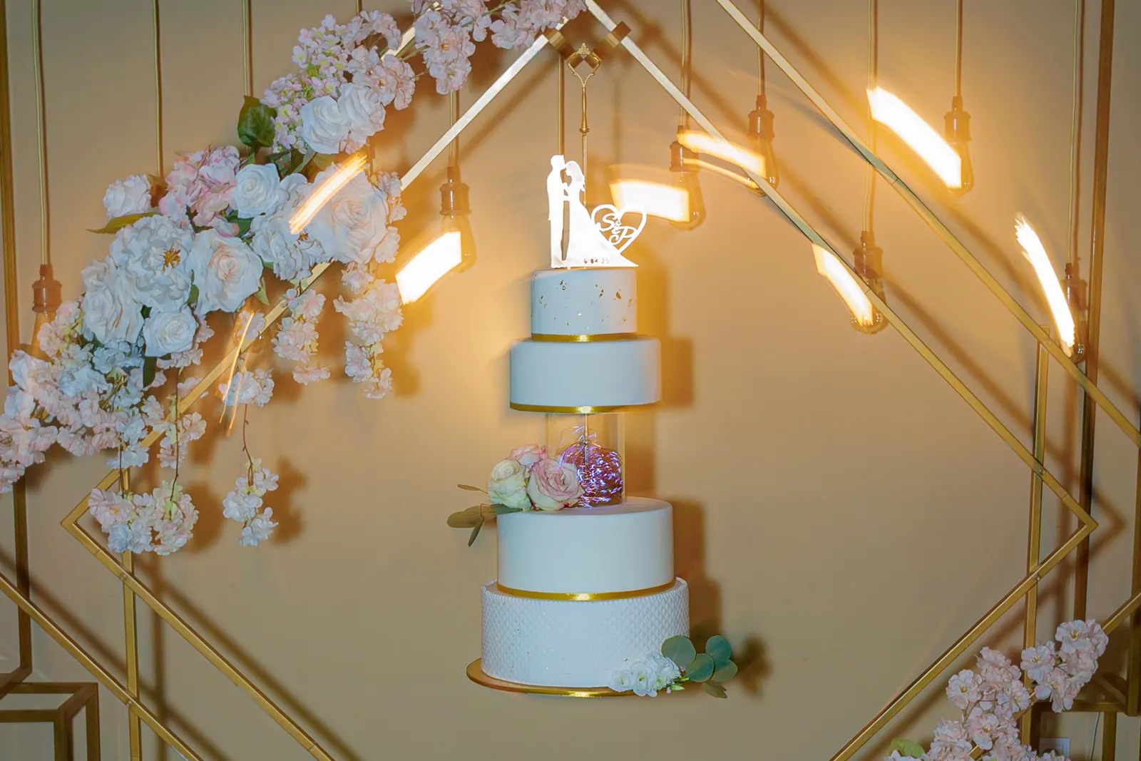 Bolo de casamento de quatro andares decorado com flores num cenário geométrico iluminado