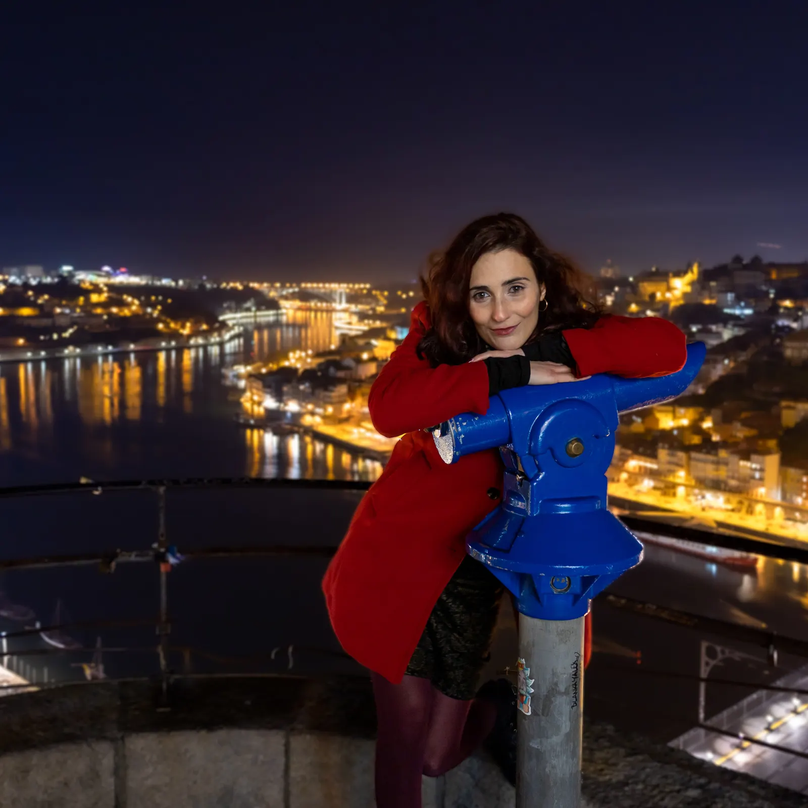Mulher de casaco vermelho apoiada num binóculo panorâmico azul à noite com a cidade iluminada ao fundo