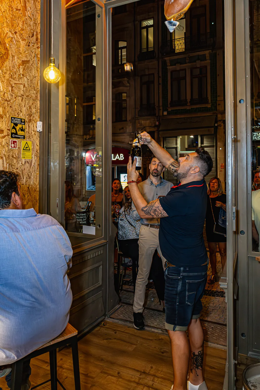 Um homem abre uma garrafa de espumante à porta de um estabelecimento comercial perante clientes atentos