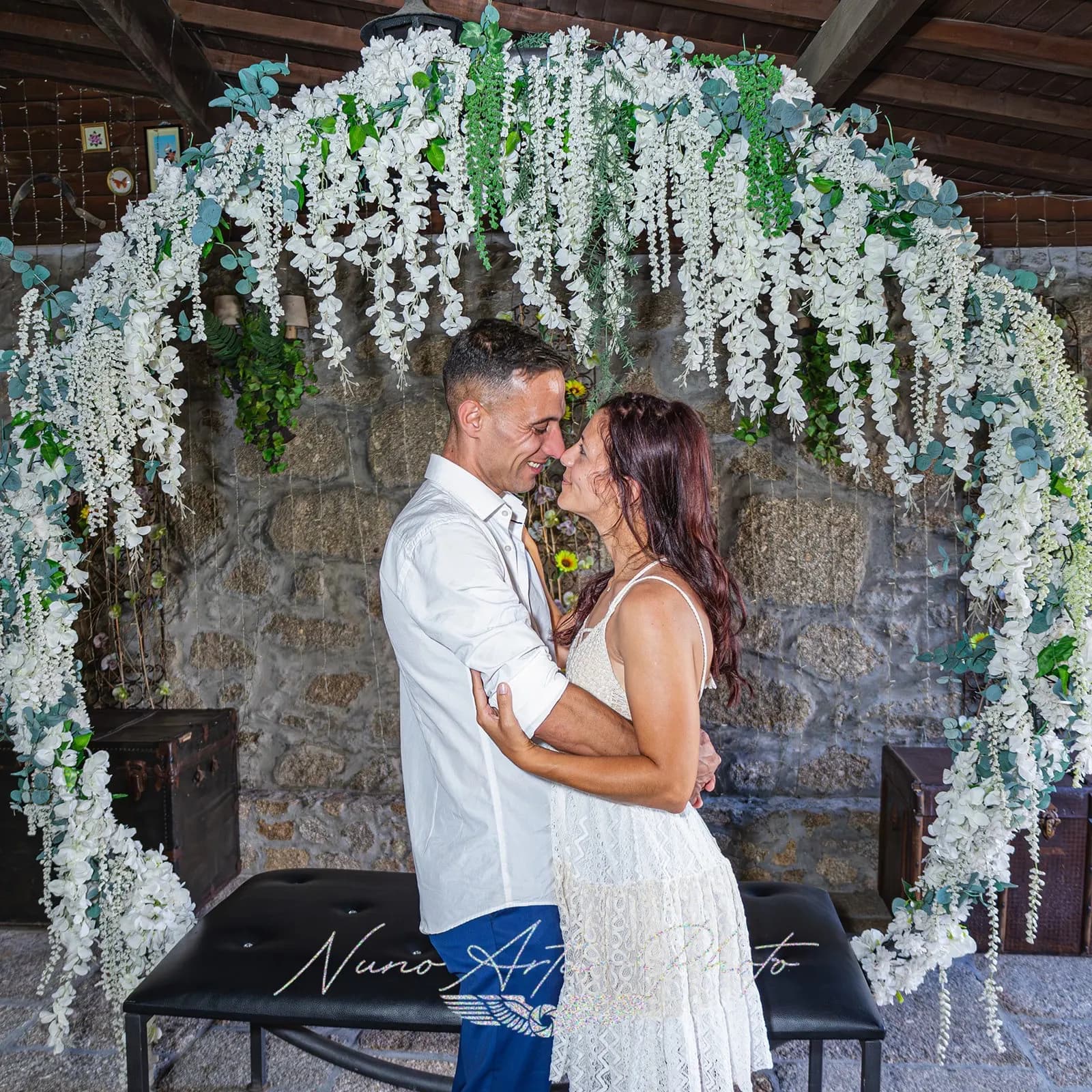 Casal abraçado sob um arco de flores brancas, diante de uma parede de pedra