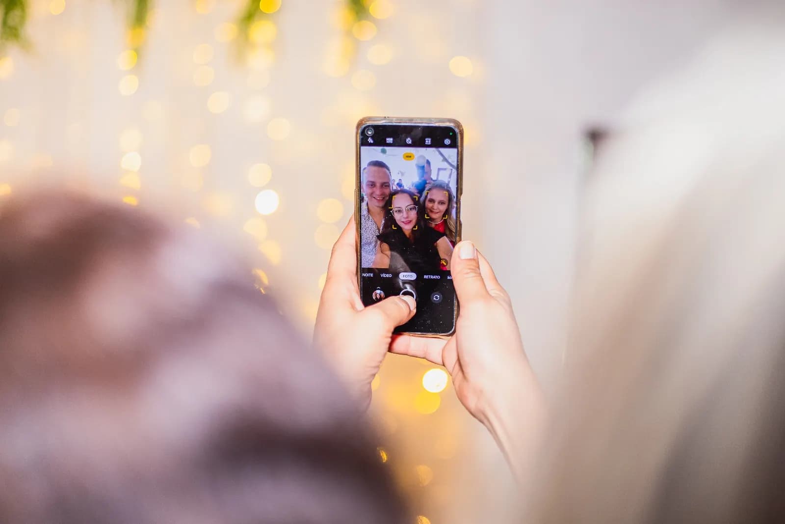 Convidados a posar no ecrã de um telemóvel, entre luzes douradas desfocadas